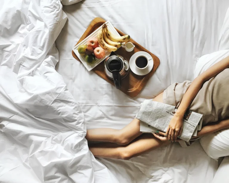 Frühstückstablett mit Kaffee und Früchten auf einem Bett, daneben eine Person mit Zeitung